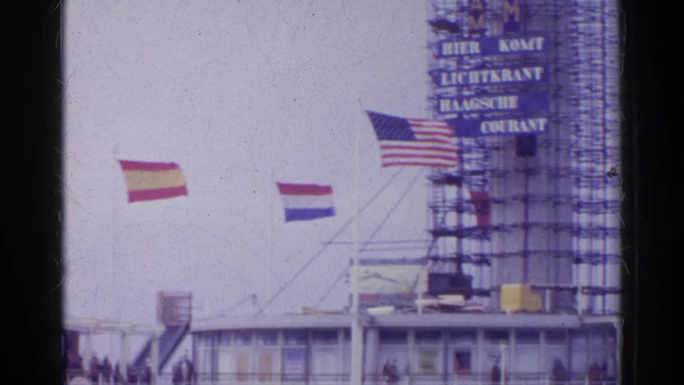 Archival home-movie frame showing several flags beside a scaffolded signboard and a low public-facing building facade.