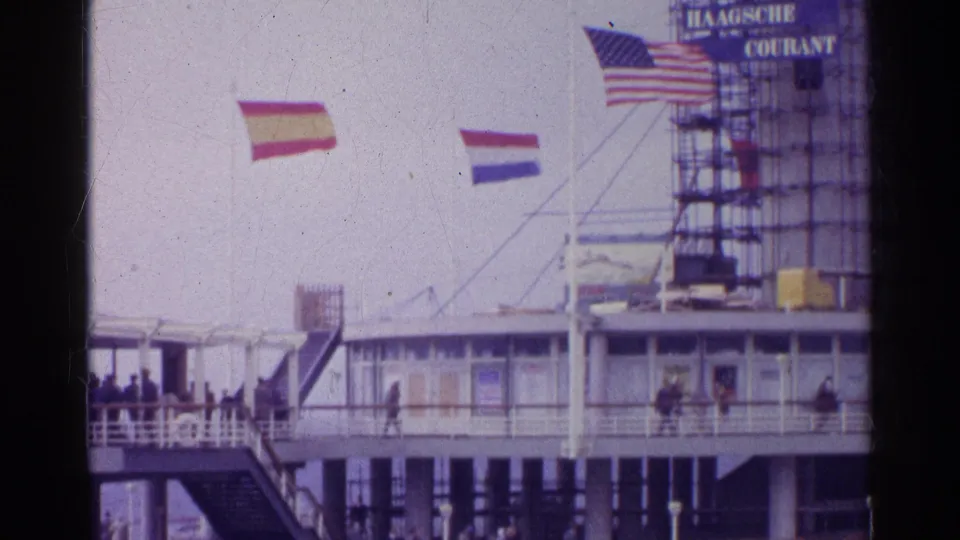 Archival home-movie frame showing flags, a scaffolded sign structure, and small figures walking along a building exterior.