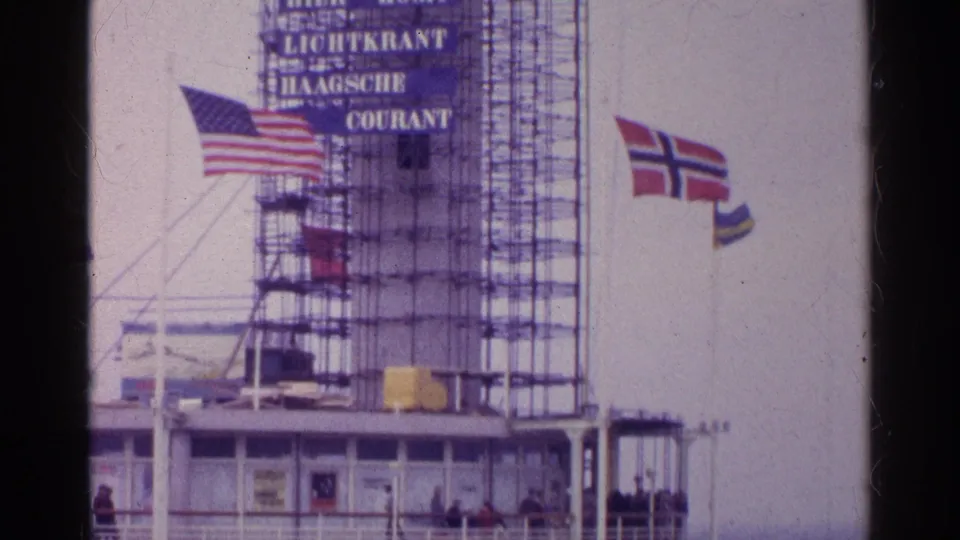 1961 8mm frame of a scaffolded ship or port structure with Haagsche Courant signage, flags, and passengers below.