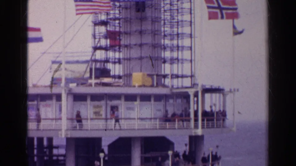 1961 ship deck scene with passengers walking below scaffolding, railings, a Dutch flag, and a Norwegian flag near the water.