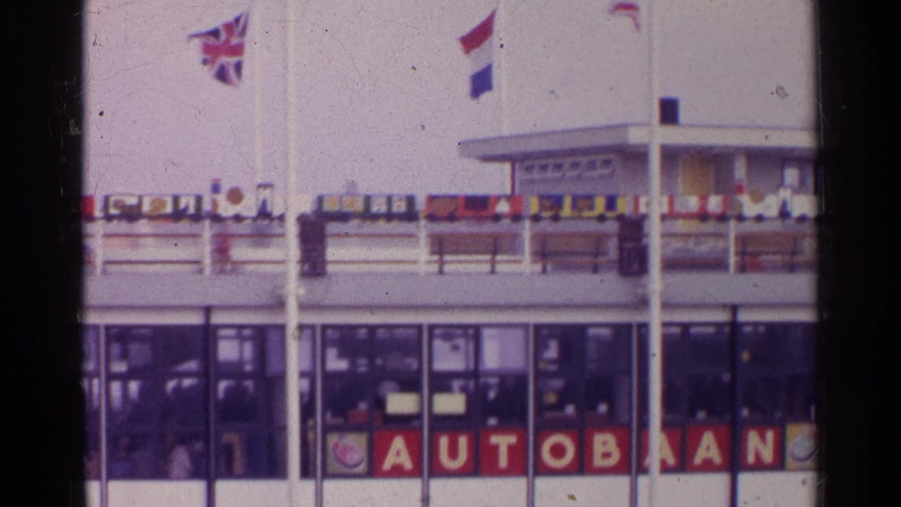 1961 Germany frame with Union Jack, Dutch and other flags flying above a ferry or terminal facade marked AUTOBAAN.