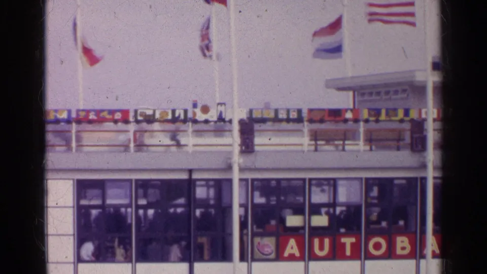1961 Germany frame showing a wider section of the AUTOBAAN-marked ferry or terminal with flags, windows, railings, and people inside.