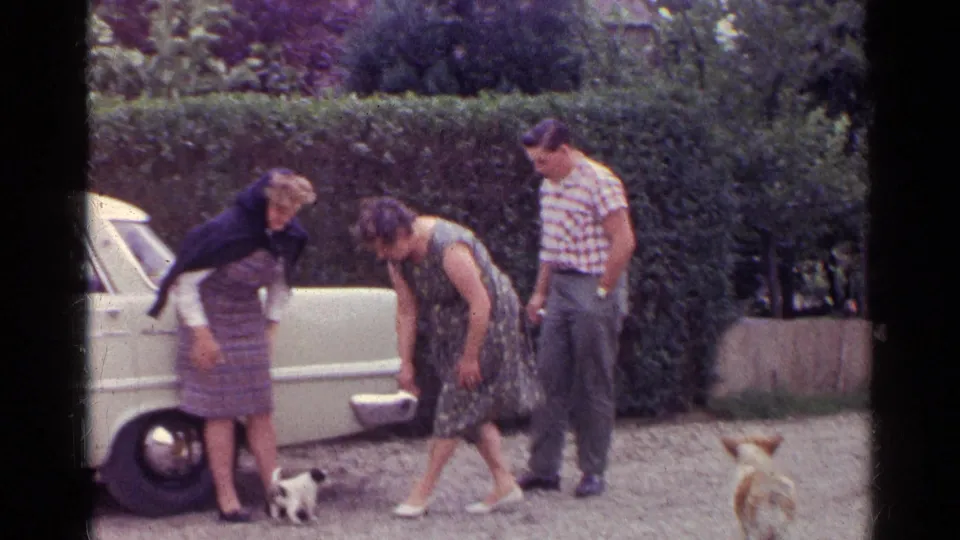Three adults stand and bend toward small dogs beside a light-colored classic car in the supplied 1961 Germany home movie.