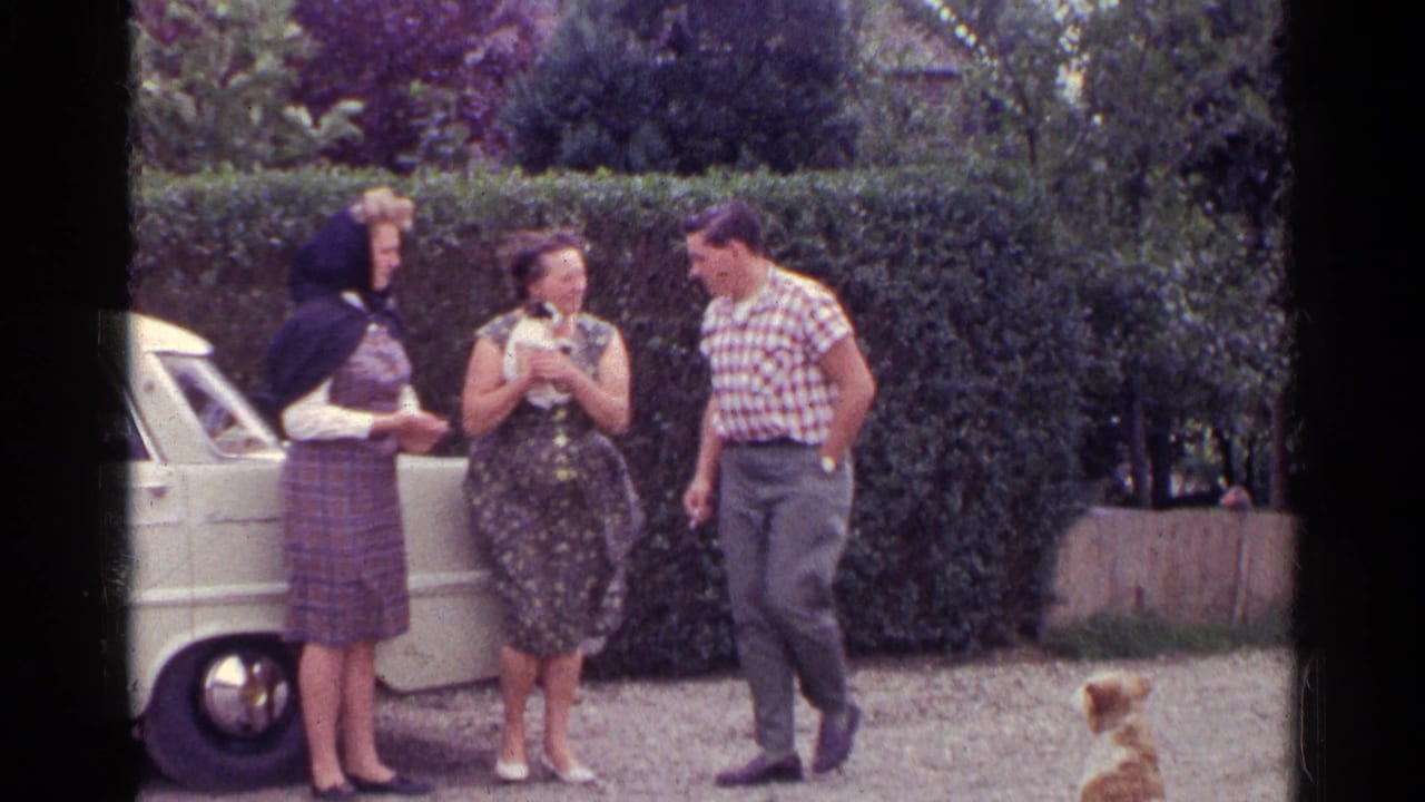 Three adults in 1961 Germany stand by a white car and focus on a small white-and-black puppy, with a tan dog nearby.