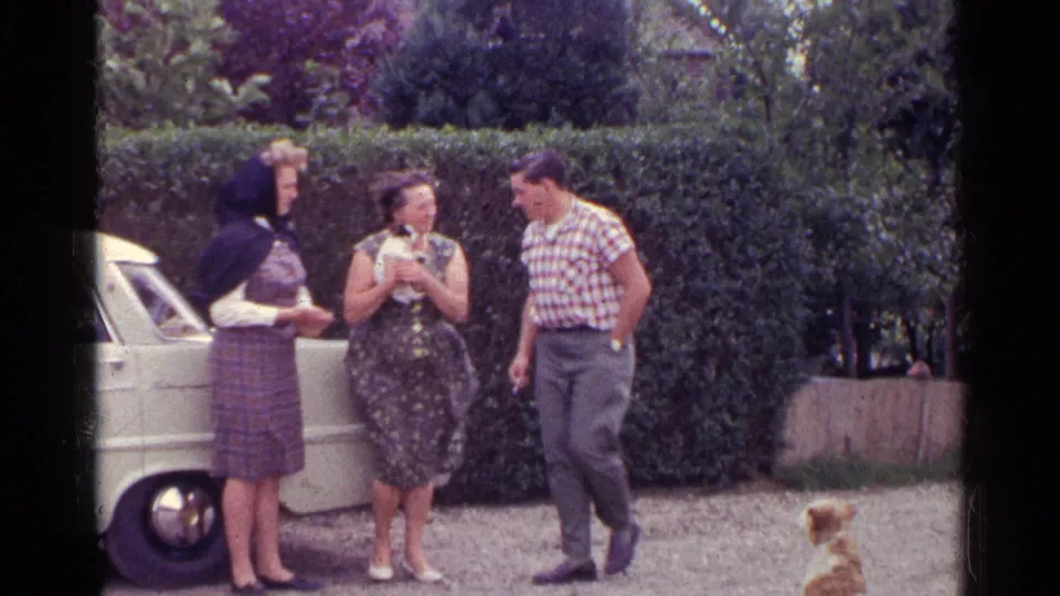 Three adults in 1961 Germany stand by a white car and focus on a small white-and-black puppy, with a tan dog nearby.