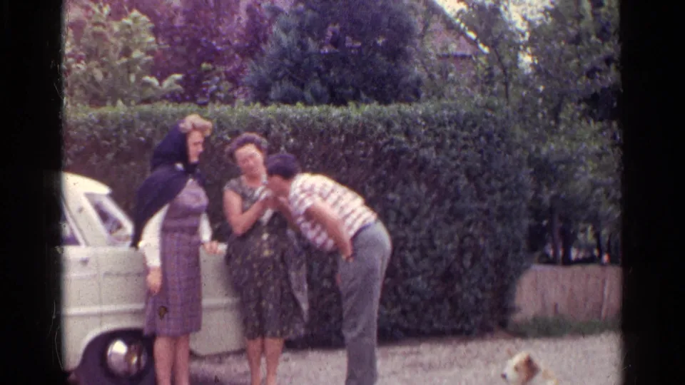 A man leans close to a small puppy held by a woman as another woman watches in an outdoor 1961 Germany home-movie scene.