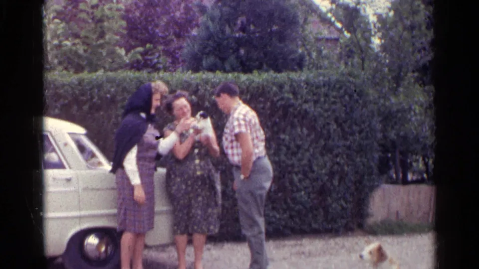 Three adults in a German 1961 home-movie scene look at a small puppy near a white car while a tan dog sits in the foreground.