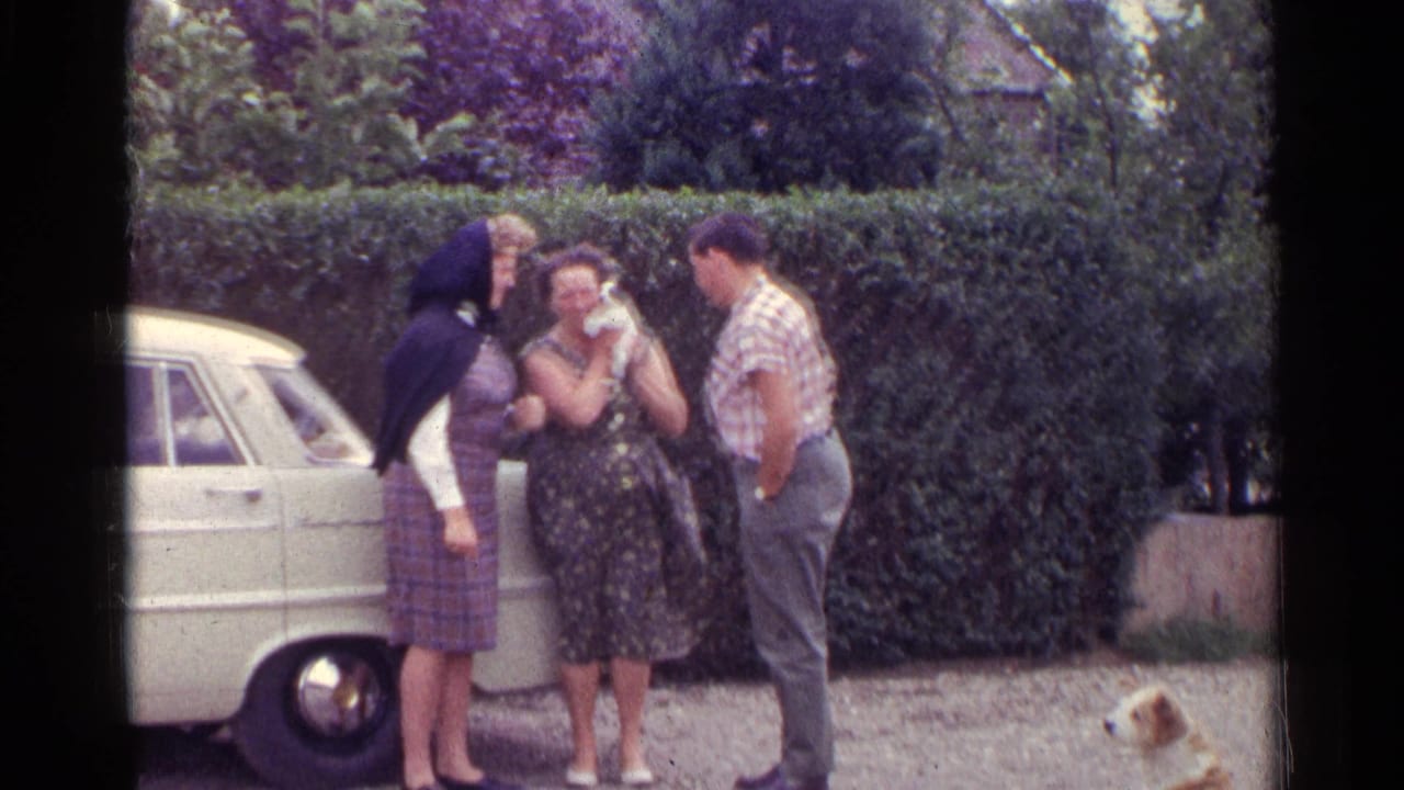 In a 1961 Germany home-movie frame, three adults gather by a white car and tall hedge while a small dog stands near them.