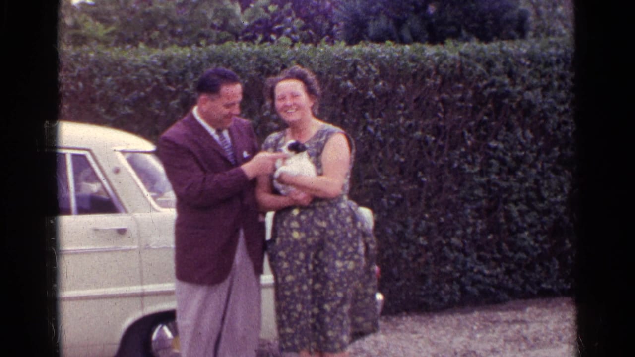 In the Germany 1961 footage, a suited man gently touches a white and black puppy held by a smiling woman near a hedge and car.