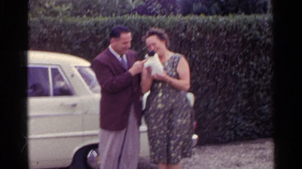 A 1961 Germany home-movie still shows a man touching or petting a small puppy as a woman holds it beside a white car.
