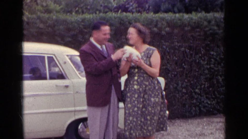 In Germany in 1961, an adult couple stand outdoors by a pale sedan as the woman holds a small black-and-white puppy.