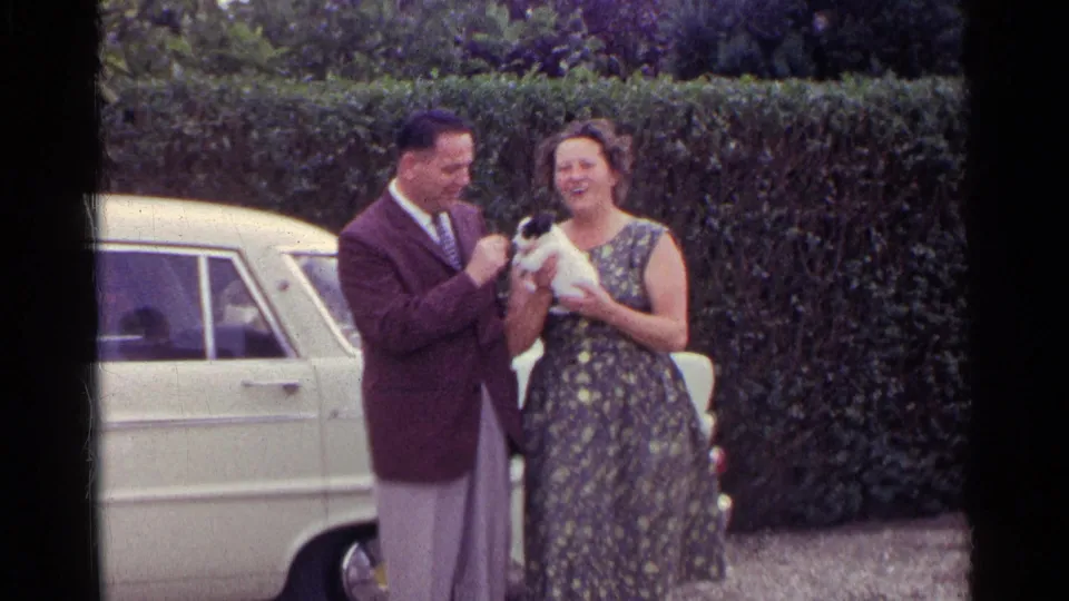 The couple smile outdoors as the woman cradles a small puppy, with a pale sedan and clipped hedge behind them.