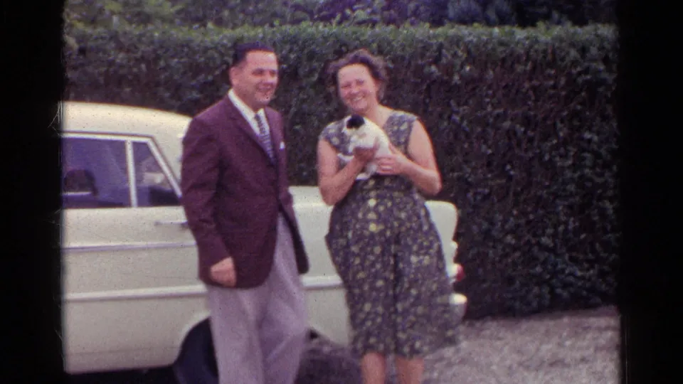 The couple stand close beside a pale sedan, smiling as the woman holds the small black-and-white puppy against her dress.
