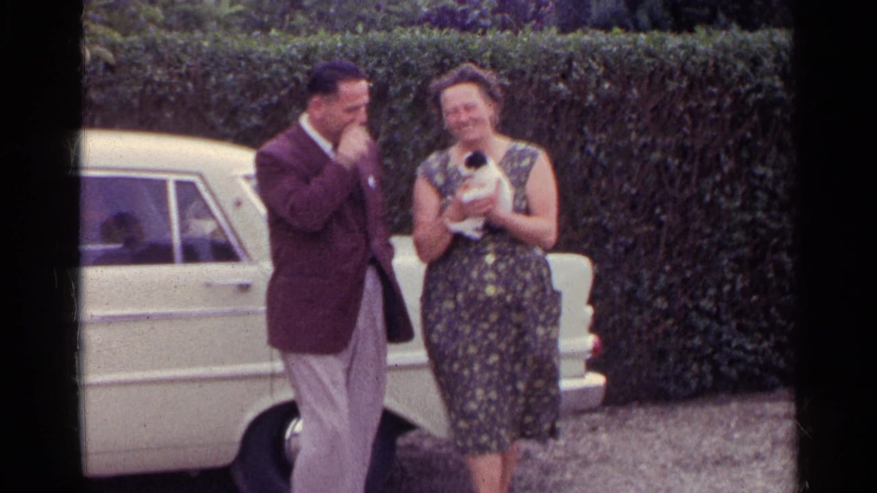 Man in suit stands beside woman holding a small kitten near a white car and hedge.