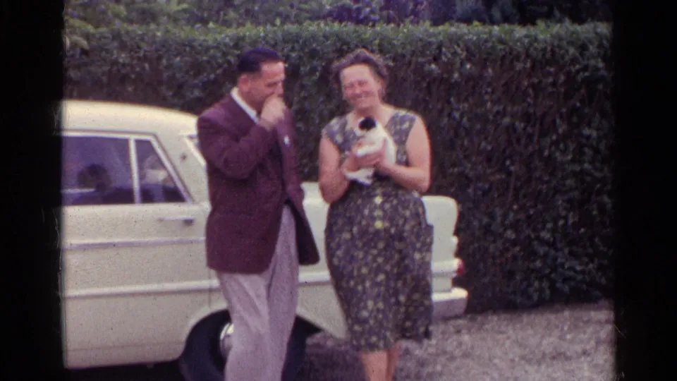 In 1961 Germany home-movie footage, a suited man smiles beside a woman in a floral dress holding a small kitten near a white car.