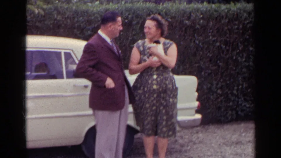 The man and woman appear to talk and smile beside a parked white car, with the woman holding a small white kitten.