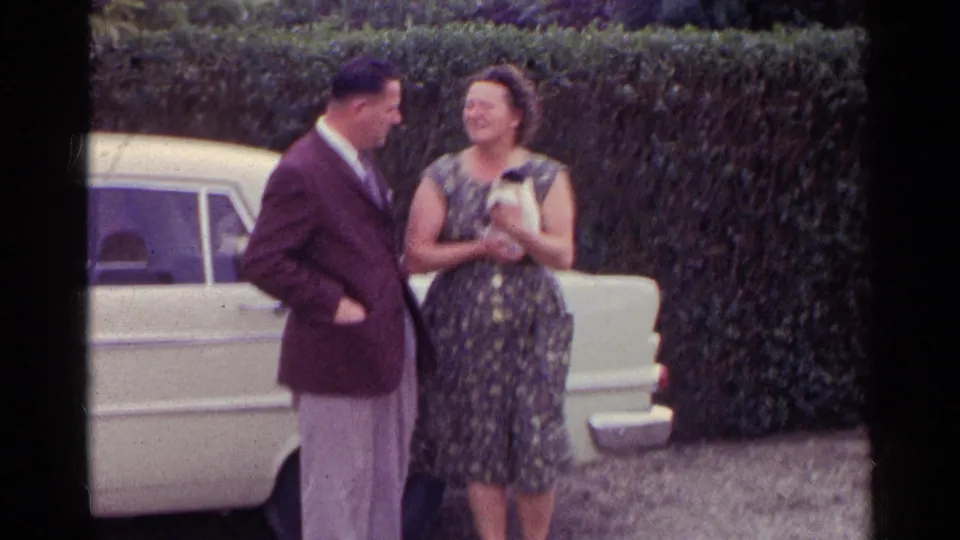 In a soft 8mm frame, the man leans toward the woman as she cradles the kitten near the white car and hedge.
