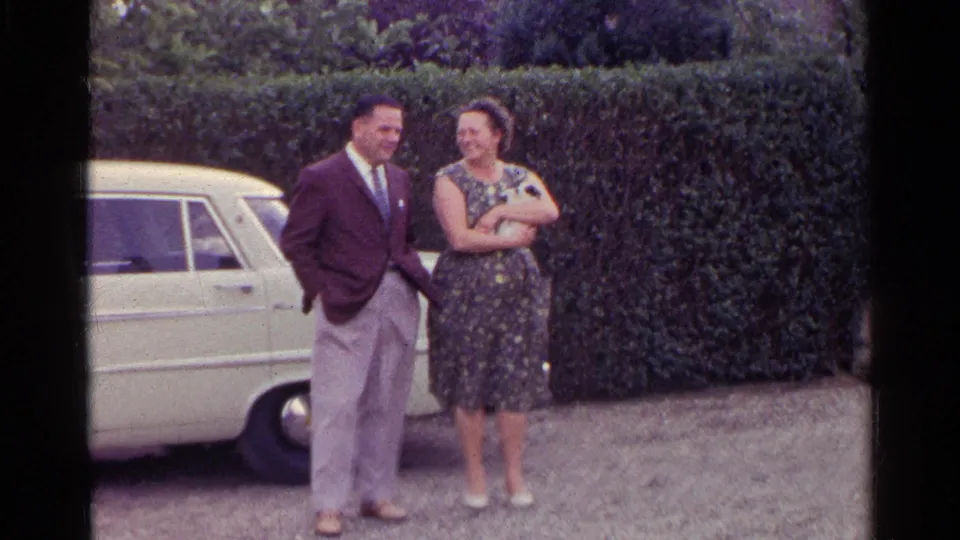 A wider 1961 Germany home-movie frame shows the couple standing beside the white car, the woman still holding the kitten against a hedge backdrop.