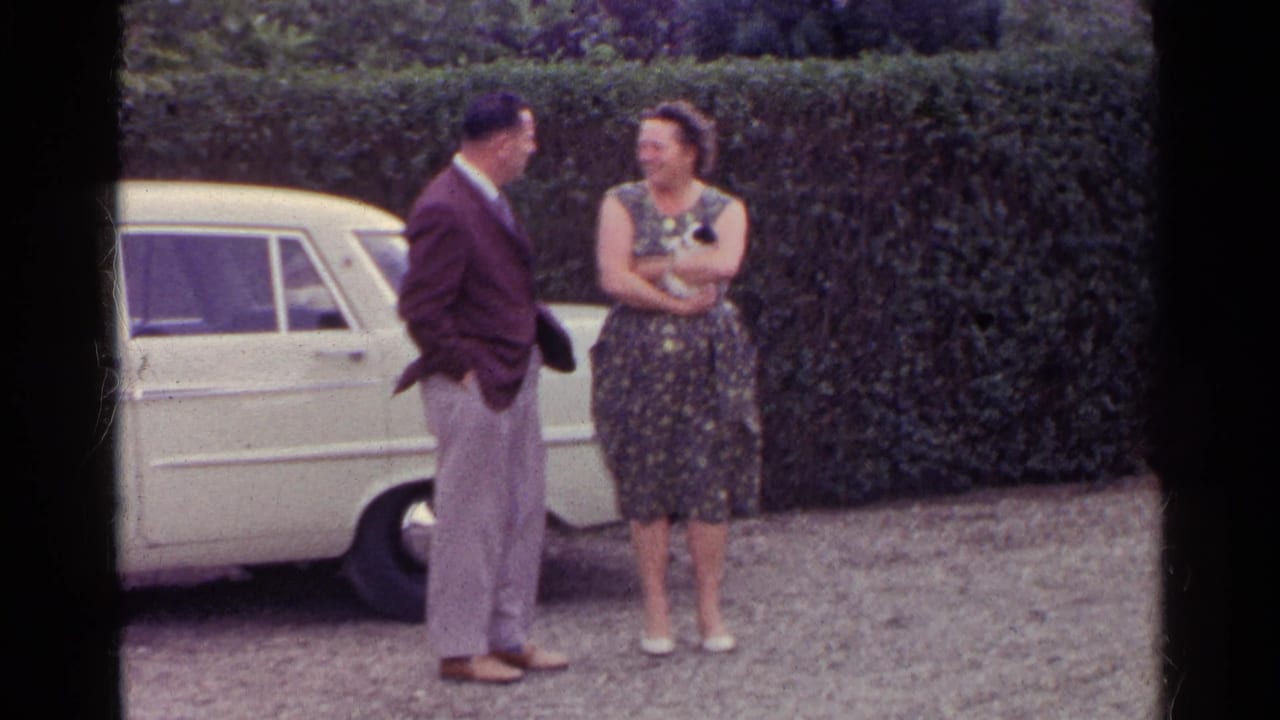 Man in maroon jacket talks with woman in floral dress beside a cream car and hedge.