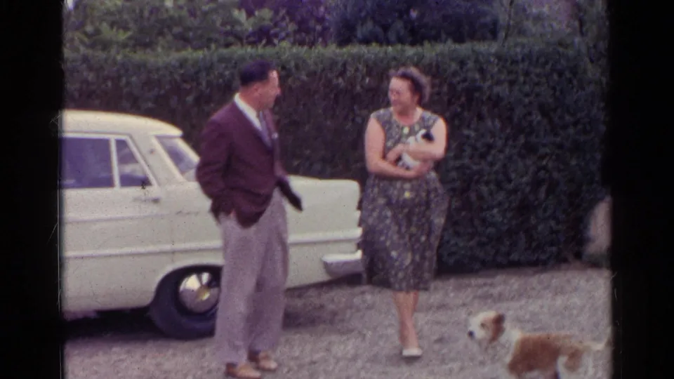 The woman holds a small white dog while a brown and white dog moves into the foreground near the adults and parked car.