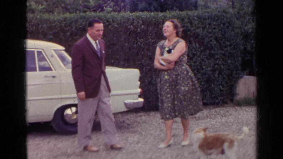 The adults appear amused in the driveway as a brown and white dog stands in front of them near the cream-colored car and hedge.