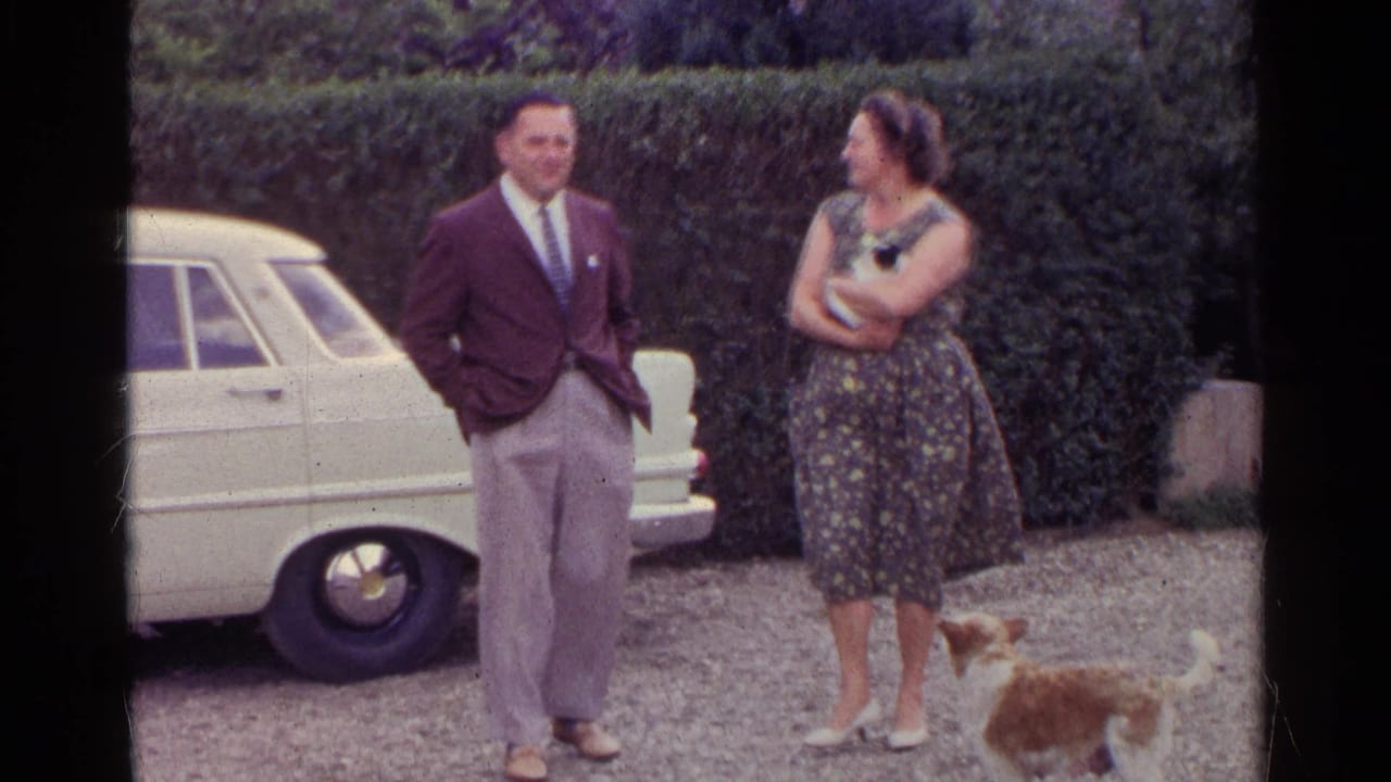1961 Germany home-movie frame of an adult man and woman talking beside a white car, with a small dog on gravel.