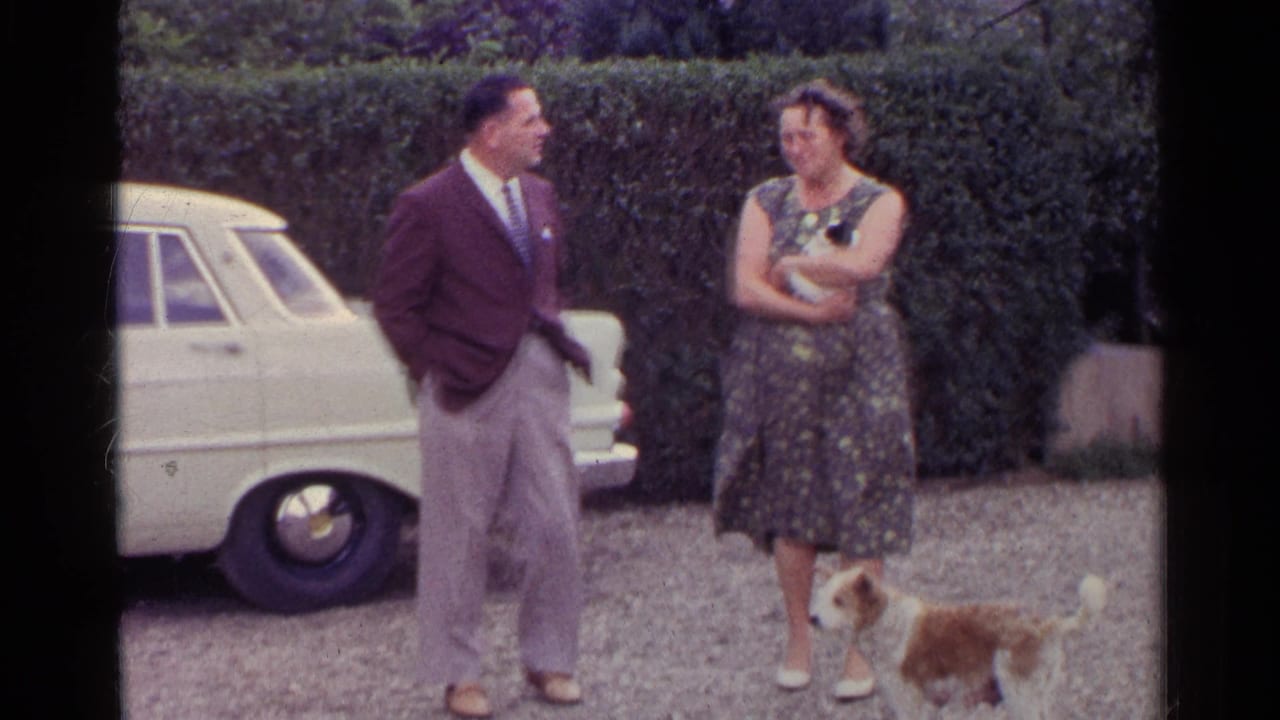 1961 Germany home-movie frame of two adults beside a white car, the woman holding an item while a small dog crosses the gravel.