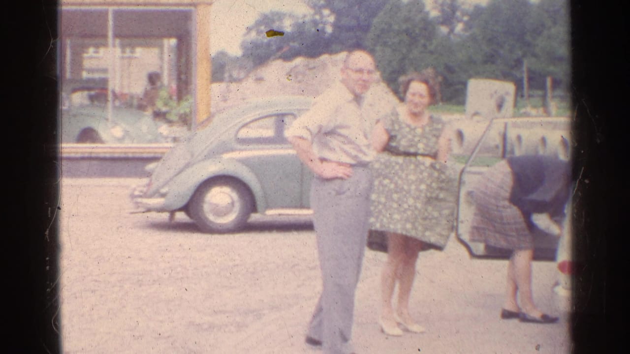 1961 Germany home-movie frame of two adults in front of a green Volkswagen Beetle near a building window.