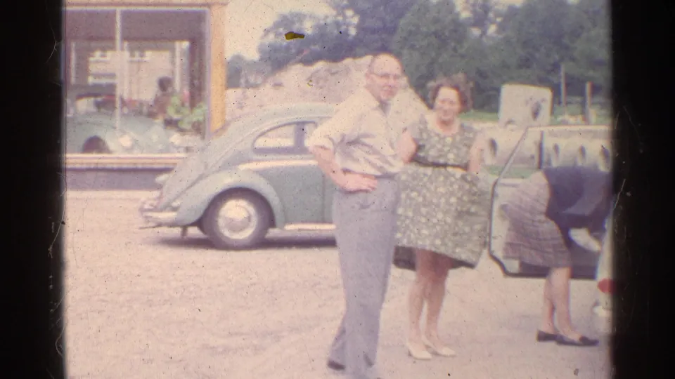1961 Germany home-movie frame of two adults in front of a green Volkswagen Beetle near a building window.