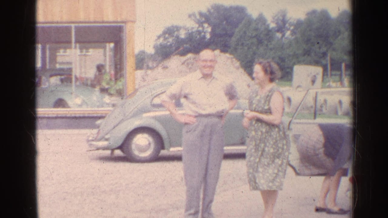 1961 Germany home-movie frame of adults beside a green Volkswagen Beetle, with a building and open car door in the background.