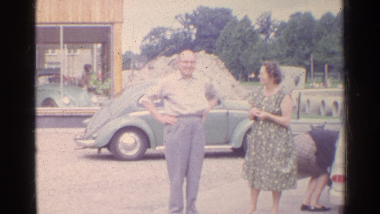 In 1961 Germany, an older man and woman stand by a green Volkswagen Beetle while facing the camera in a home-movie frame.