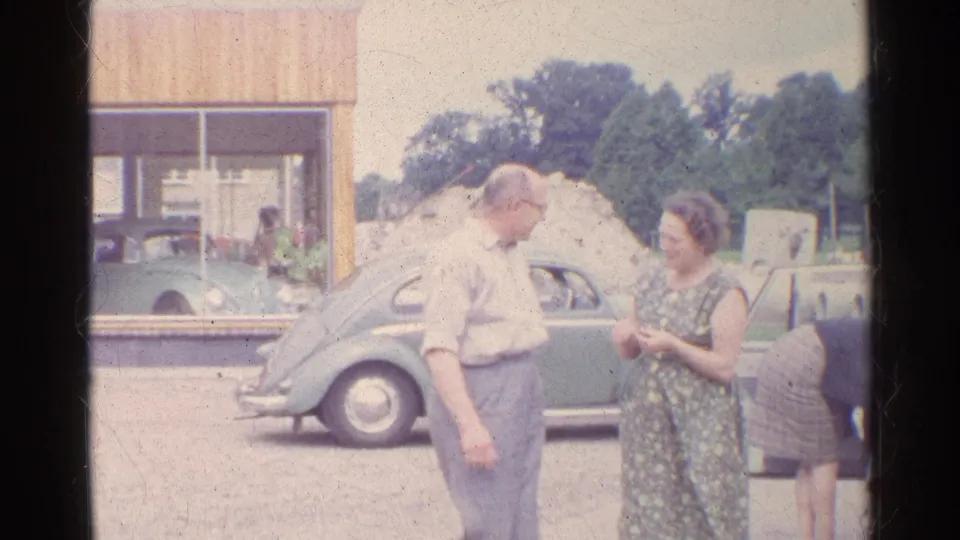 The older man turns to speak with the woman beside the green Volkswagen Beetle, with a building and trees behind them.