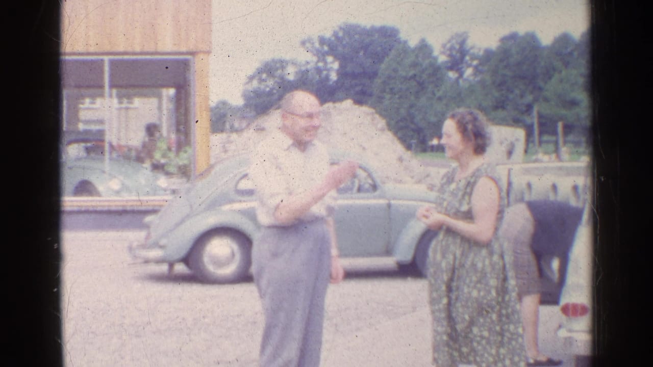 In a soft 8mm frame, the older man gestures toward the woman as they converse beside the parked Volkswagen Beetle.