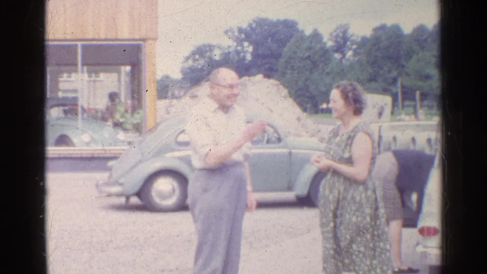 In a soft 8mm frame, the older man gestures toward the woman as they converse beside the parked Volkswagen Beetle.