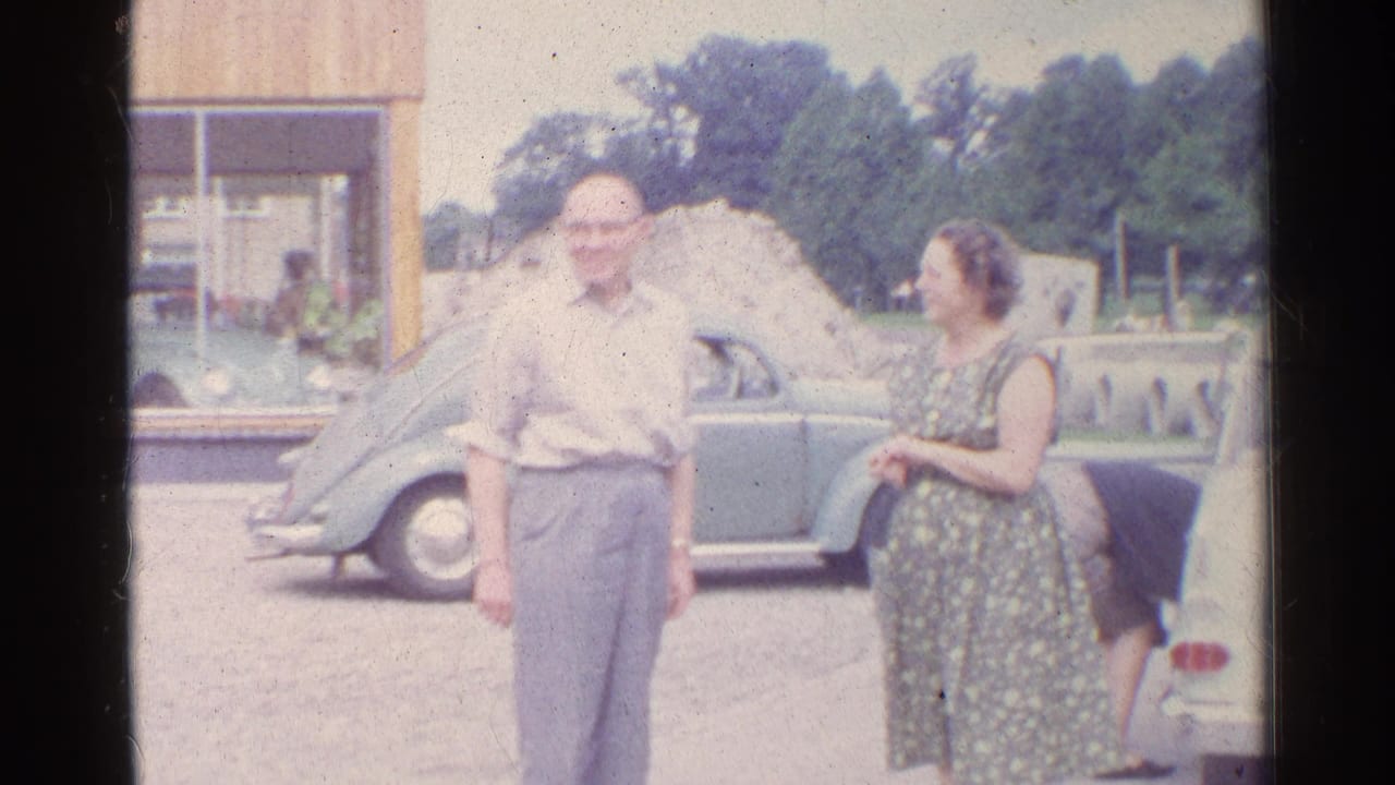 The older man smiles toward the camera as the woman stands near him and another person is partly visible at the car.