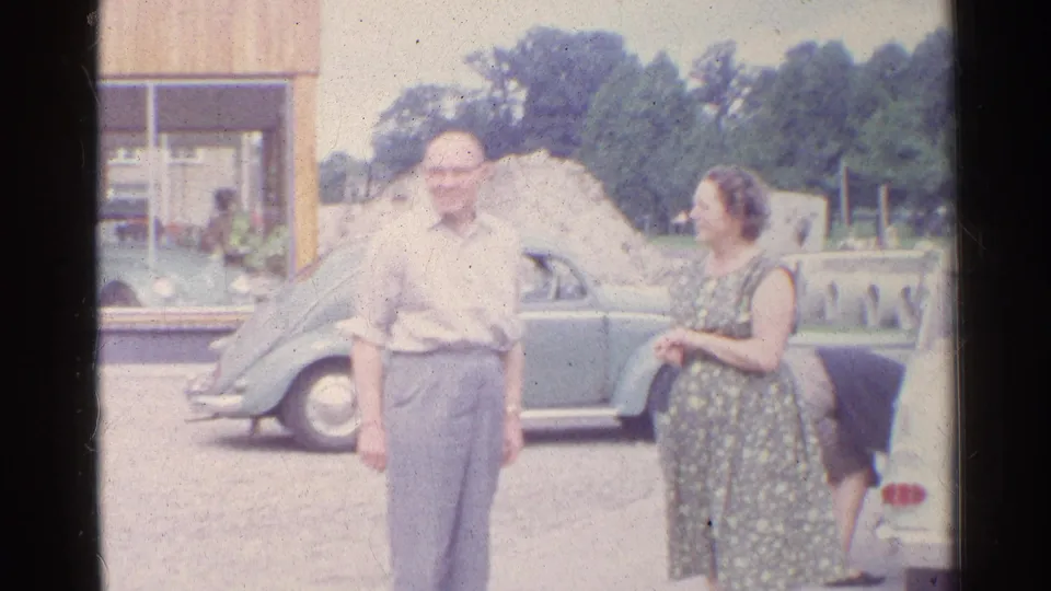 The older man smiles toward the camera as the woman stands near him and another person is partly visible at the car.