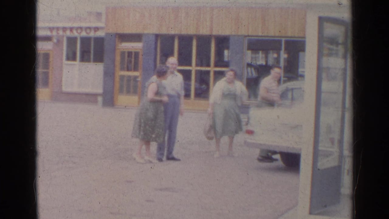 Four adults stand near a light-colored car outside storefronts with a partial VERKOOP sign.