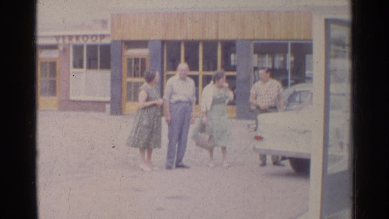 Two women and two men stand near the rear of a light-colored car, with storefront glass and a partial sign behind them.