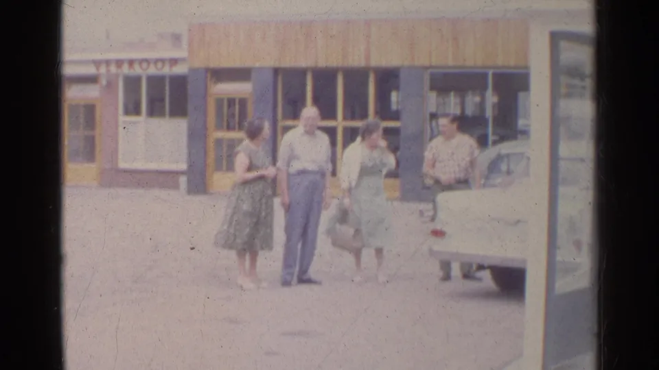 Two women and two men stand near the rear of a light-colored car, with storefront glass and a partial sign behind them.