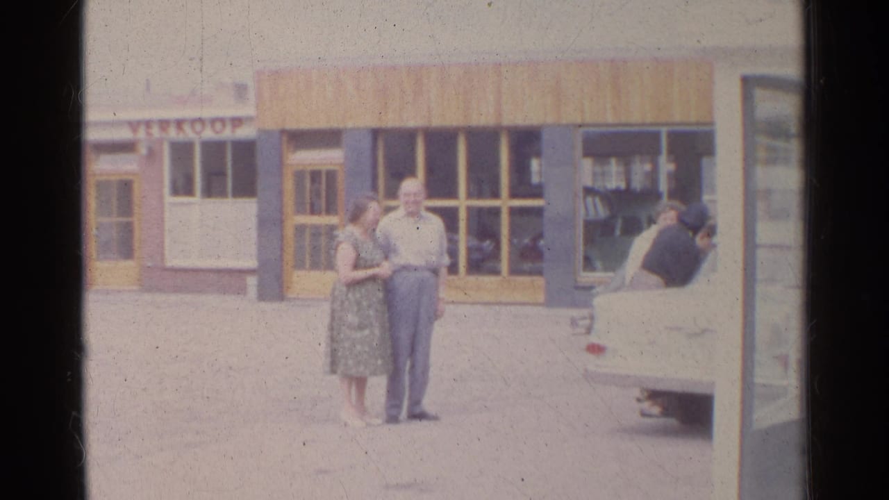 1961 Germany frame of a woman in a patterned dress and a man in light clothing standing by shop windows near a white car.