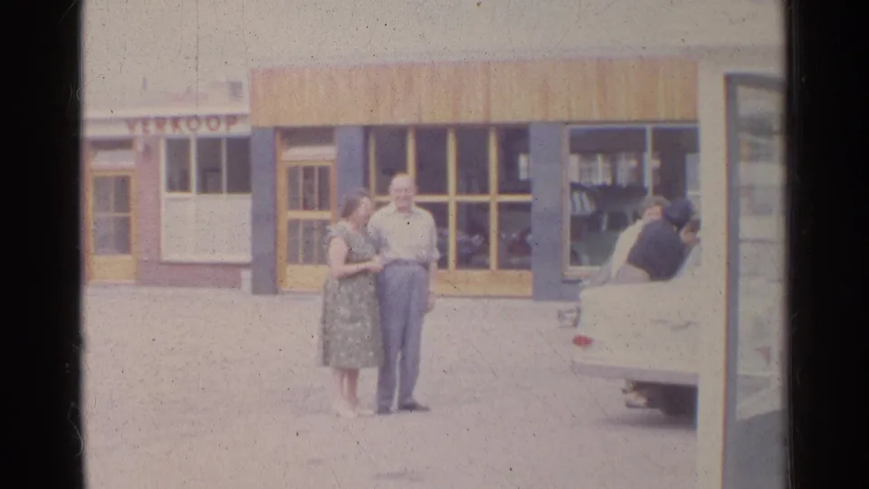 1961 Germany frame of a woman in a patterned dress and a man in light clothing standing by shop windows near a white car.
