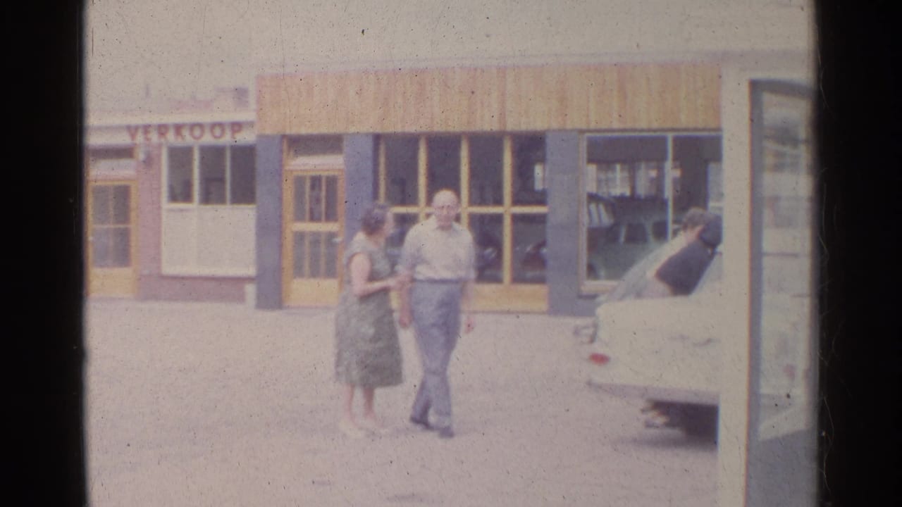 Home-movie frame from 1961 Germany showing a couple moving away from a storefront with a white car at right.