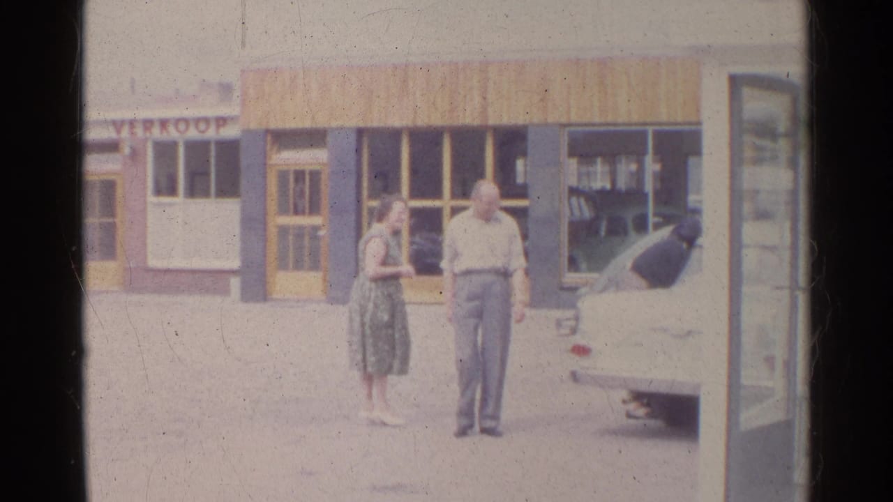 Two adults stand near a white car outside a storefront with large windows.