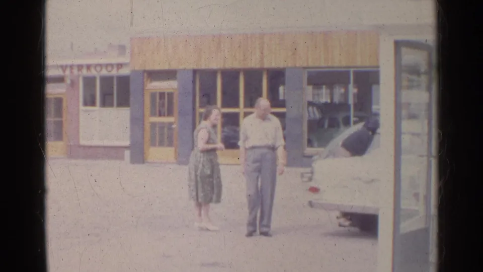 1961 Germany home-movie frame showing a man and woman conversing beside a white sedan outside a shopfront.