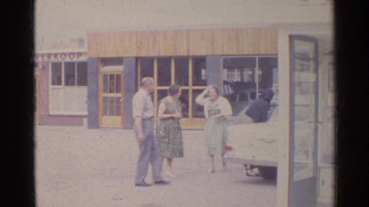 1961 Germany street scene with three adults talking beside a white sedan in front of a shopfront.