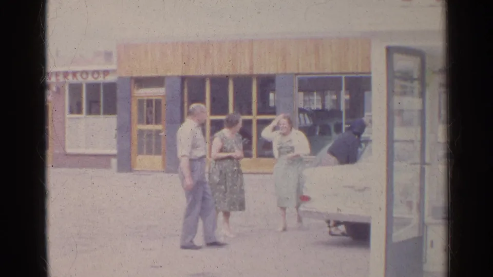 1961 Germany street scene with three adults talking beside a white sedan in front of a shopfront.