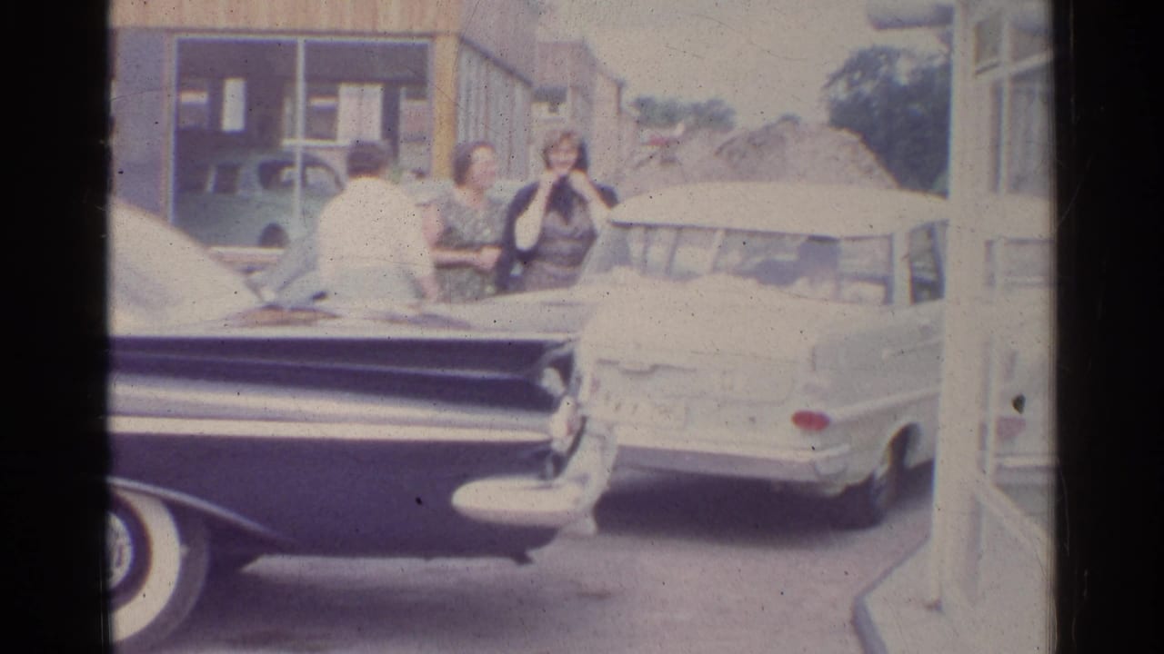 1961 Germany street view with a dark tailfin car, white sedan, and adults near storefront buildings.