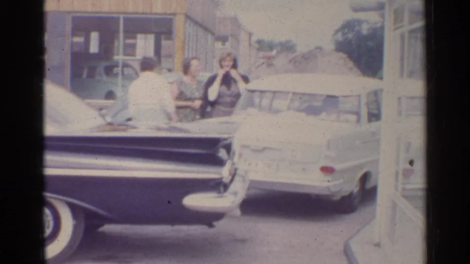 1961 Germany street view with a dark tailfin car, white sedan, and adults near storefront buildings.