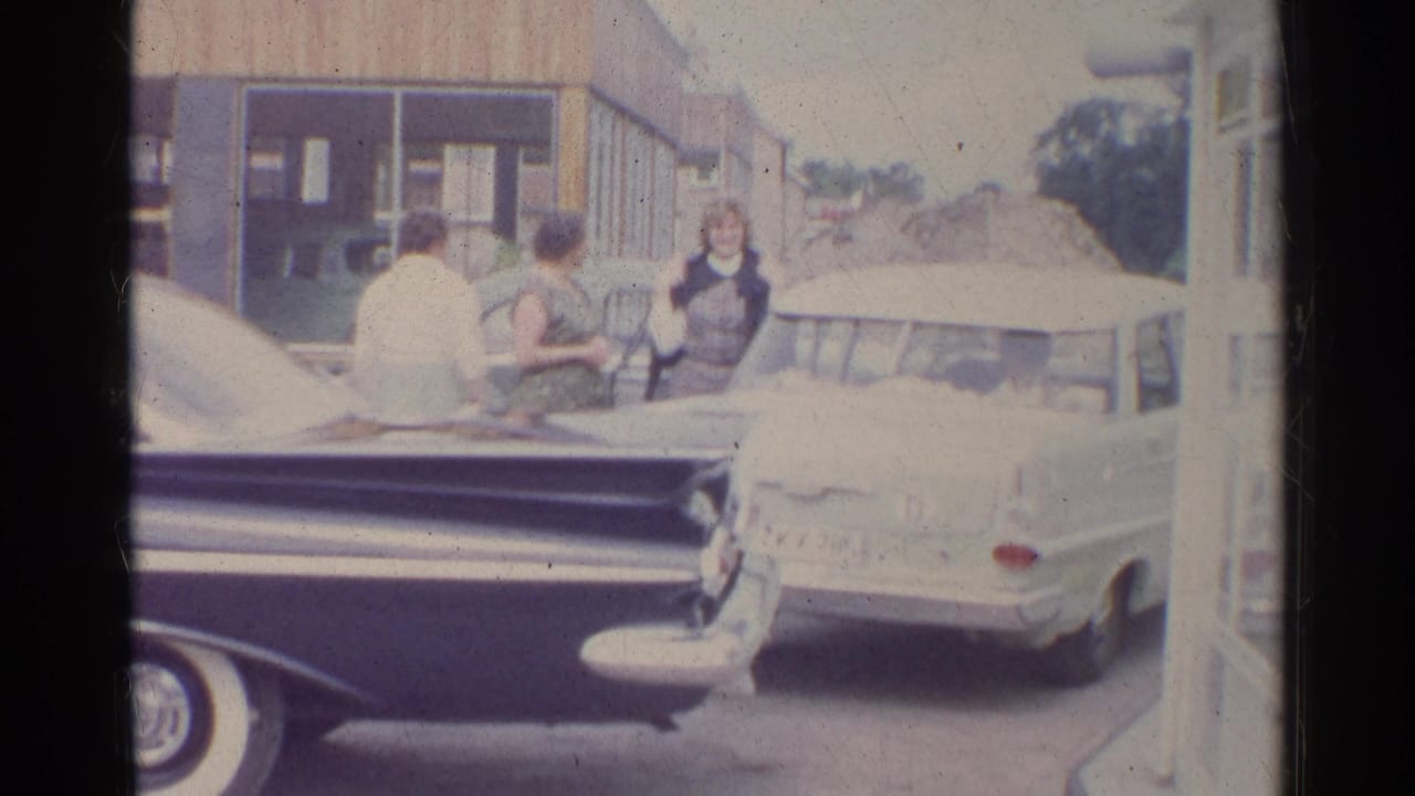 Three women stand beside parked cars outside a service-station building.