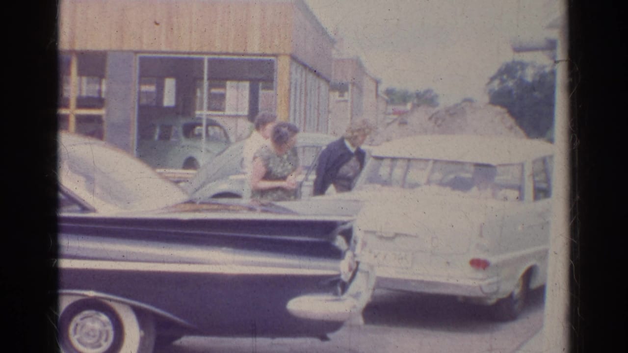 A woman bends slightly near the parked cars as another stands beside her at the 1961 German service-station scene.